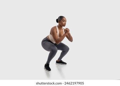 A focused African American woman athlete is engaged in a strength-building exercise routine, demonstrating perfect form while doing squats on white background - Powered by Shutterstock