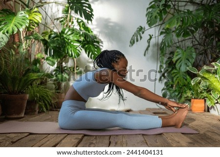 Focused african american woman enhances body flexibility as reaches for toes, stretching arms towards feet sitting on fit mat in studio with indoor plants. Athletic black female enjoy home fitness.