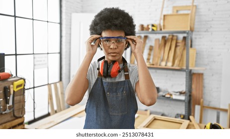 A focused african american woman wearing safety goggles and headphones in a well-lit carpentry studio. - Powered by Shutterstock