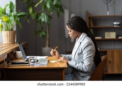 Focused African Female Office Assistant With Braided Hairstyle Writing Something Down On Sticky Note While Sitting In Front Of Laptop, Black Woman Freelancer Planning Out Day While Working Remotely