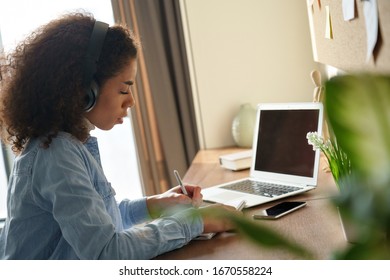 Focused african female student wears headphones elearning studying alone at home office desk. Millennial mixed race teen girl listening audio podcast e learning english language concept making notes.
