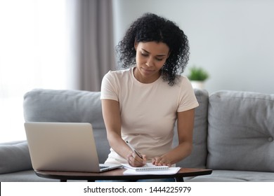 Focused african girl student study online at home making notes in notebook sit at home office desk with laptop, young black woman freelancer writing down information working distantly or e learning