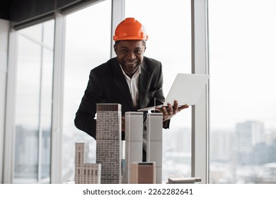 Focused african inspector using laptop inspecting skyscraper model in architectural modern office with panoramic city view on architectural bureau. Handsome man wearing suit and hardhat.