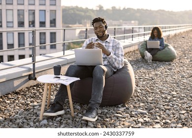 Focused African male wearing denim shirt and headset holding online conference over portable computer on rooftop, female colleague working on blurred background. - Powered by Shutterstock