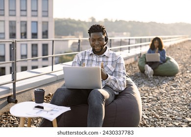 Focused African male wearing denim shirt and headset holding online conference over portable computer on rooftop, female colleague working on blurred background. - Powered by Shutterstock