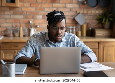 Focused African Student Guy Studying From Home, Watching Learning Webinar On Laptop, Attending Virtual Class. Serious Distance Employee, Business Professional Working At Computer In Kitchen