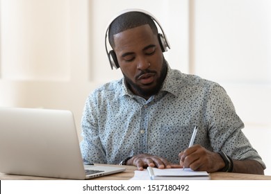 Focused African Student Sit At Desk Wearing Wireless Headphones Learning Preparing For Seminar Or Exam, Guy Interpreter Hears Audio Writing Down Translation, Online Lecture Course E-learning Concept