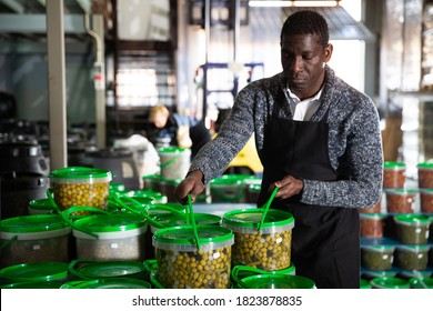 Focused African-American Man Working In Food Producing Factory, Arranging Plastic Buckets With Pickled Olives