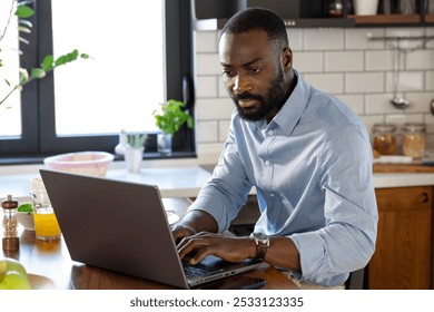 A focused African-American man works on his laptop at a kitchen counter. The modern kitchen is well-lit, with fresh fruit and a glass of juice nearby, creating a productive atmosphere. - Powered by Shutterstock