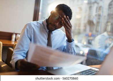 Focused African-American office manager sitting at cafe table with laptop, reading important documents with puzzled expression and holding head with his hand. - Powered by Shutterstock