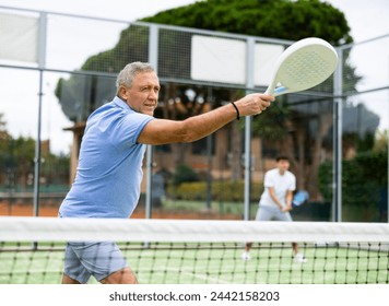 Focused aged man playing friendly paddleball match on outdoor summer court. Senior people sports concept.. - Powered by Shutterstock