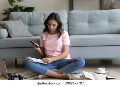 Focused Arabian woman writing taking notes, holding smartphone, sitting on floor with laptop and books, young female student watching webinar, involved in online course, preparing to exam at home