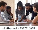 Focused arican american millennial female team leader leaned over table, writing down project ideas, editing documentation at brainstorming business meeting with diverse partners or clients at office.