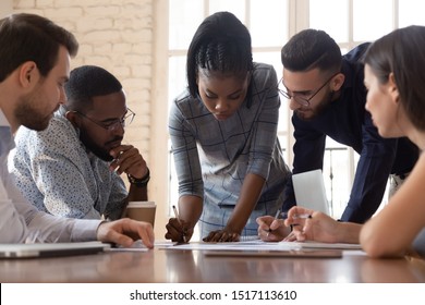 Focused arican american millennial female team leader leaned over table, writing down project ideas, editing documentation at brainstorming business meeting with diverse partners or clients at office. - Shutterstock ID 1517113610