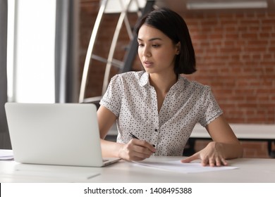 Focused Asian Business Woman Working Studying Online In Office Looking At Laptop Making Notes, Serious Japanese Employee Or Student Watching Webinar Writing Information In Notebook Sitting At Desk