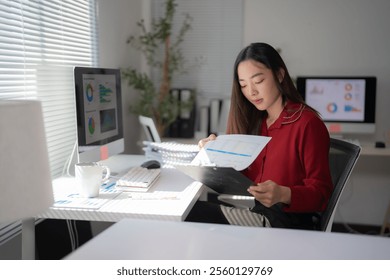 Focused asian businesswoman reviewing financial reports and data analysis on clipboard and computer, working diligently in modern office workspace - Powered by Shutterstock