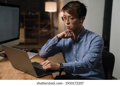 Focused Asian Guy In Earphones Working With Laptop While Sitting At Table In Office