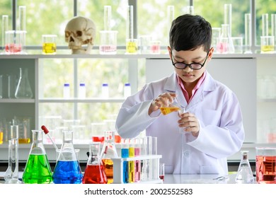 Focused Asian Kid In White Coat And Glasses Pouring Yellow Liquid Into Flask While Conducting Experiment With Colorful Reagents During Chemistry Research In Laboratory.