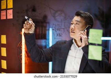 Focused Asian Male Ceo Manager In Formal Wear Writing On Glass Board With Colorful Sticky Notes And Talking Phone, Working With Project In Modern Night Office. Creative Thinking, Startup Plan Concept