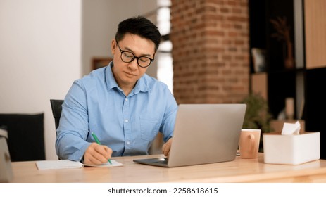 Focused asian mature man using laptop, working remotely and taking notes, having remote meeting from home, panorama. Middle aged man studying online, writing down info - Powered by Shutterstock