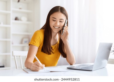 Focused asian woman multitasking with phone and notepad beside an open laptop, working at home office - Powered by Shutterstock