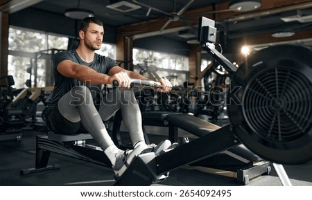 Focused athletic man using a rowing machine during workout at a bright, spacious gym. Concept: endurance, cardio, fitness, health, discipline.