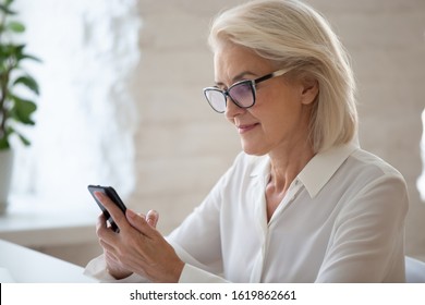 Focused attractive middle-aged businesswoman in glasses seated at workplace desk holding smart phone read latest news, check e-mail, using business application, typing message solve issues distantly
