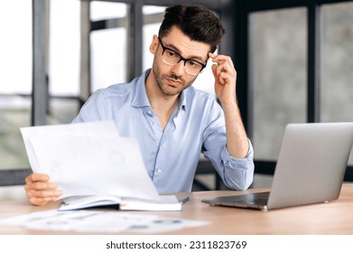 Focused attractive smart successful young caucasian businessman, top manager or broker wearing glasses, sitting at workplace, looking through business documents, thinking over a business strategy - Shutterstock ID 2311823769