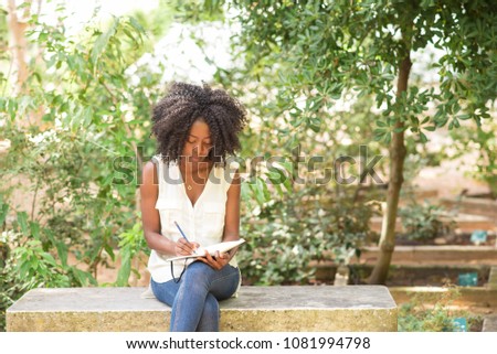 Focused attractive young black woman making notes in park. Girl sitting on concrete bench and writing with plants in background. Freelancing concept. Front view.