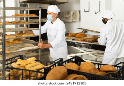 Focused baker in uniform and protective mask for disease prevention carrying tray rack trolley with bread in kitchen of bakery - Powered by Shutterstock