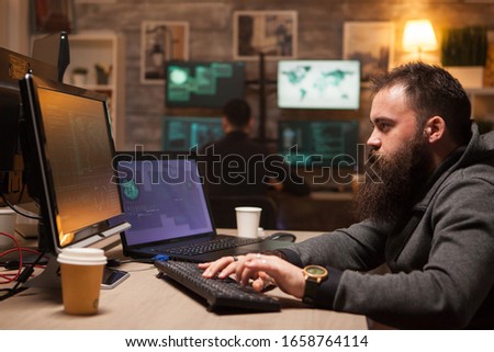 Focused bearded hacker after drinking a lot of coffee to stay awake. Young cyber criminal in the background.