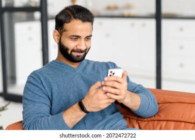 Focused bearded Indian man lying on the sofa at home and holding smartphone, Hispanic guy reading news online, spend leisure time in social networks, dark-haired arab male texting and chatting online