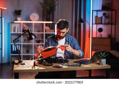 Focused Bearded Man In Denim Shirt Sitting At Table And Repairing Remote Controlled Car. Caucasian Male Trying To Fix Favorite Child Toy At Home.