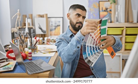 A focused bearded man examines a color swatch in a creative studio setting, surrounded by art supplies and a laptop. - Powered by Shutterstock