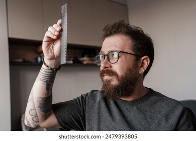 A focused bearded man with glasses examining a strip of film negatives, revealing a blend of photography skills and modern interiors. - Powered by Shutterstock