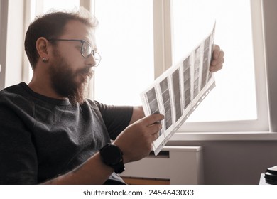 Focused bearded man with glasses looking at film negatives in bright natural light beside a window. - Powered by Shutterstock