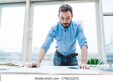 Focused bearded man in shirt thinking while standing with pencil by table with papers by window in light modern office