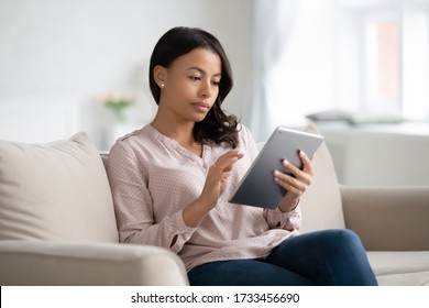 Focused Beautiful African Ethnicity Woman Sitting On Couch In Living Room Holding Tablet Gadget, Freelancer Working From Home, Housewife Choose Goods On-line Surf Websites Stores, Read News Concept