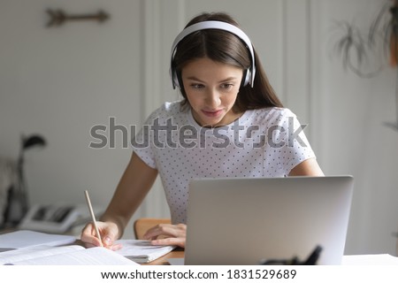 Focused beautiful young woman student in headphones looking at computer screen, listening to educational lecture webinar or video conference, making notes in copybook, distant education concept.