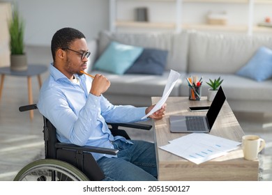 Focused Black Disabled Man In Wheelchair Working With Documents, Using Laptop At Home Office. Handicapped Afro Man Sitting At Desk With Computer, Checking Financial Reports