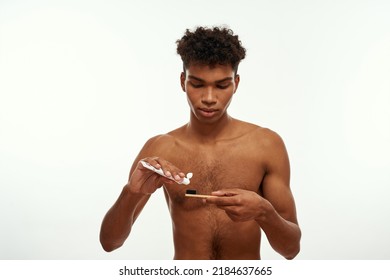 Focused Black Guy Pouring Toothpaste On Toothbrush Before Brushing Teeth. Young Brunette Curly Man With Naked Torso. Dental Care And Hygiene. Isolated On White Background. Studio Shoot. Copy Space