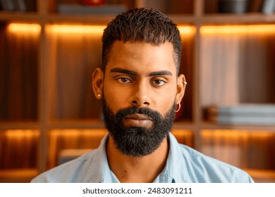 Focused black man with a beard posing indoors, wearing a light blue shirt. Serious young male with stylish haircut and piercing, standing against a warm, wooden background