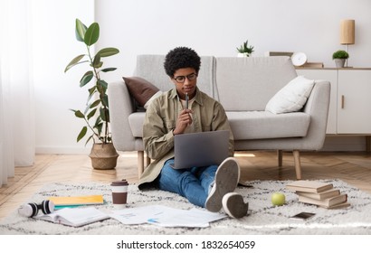 Focused black teen guy sitting on floor with study materials and laptop, at home. African American teenager studying remotely, attending online lecture or seminar, communicating with college teacher