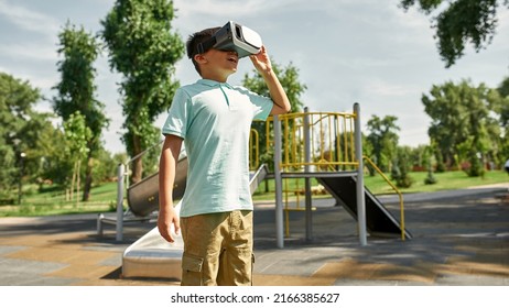 Focused Boy With Open Mouth Watching Virtual Reality Headset Outdoors. Male Child Of Digital Generation Alpha. Modern Childhood Lifestyle. Gadget Addiction. Playground In Park. Sunny Summer Day