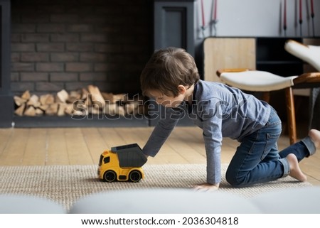 Focused boy playing with toys on heating floor at home, wheeling plastic car, trailer truck on carpet. Kid involved in game activity, improving imagination, creative skills. Childhood in safe house