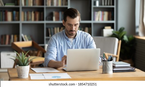 Focused Business Man Sit At Workplace Desk At Home Office Cozy Room Working On Laptop, Make Analysis Sales Stats Results, Learn Graphs And Making Financial Forecasts. Busy Workday, Modern Tech Concept