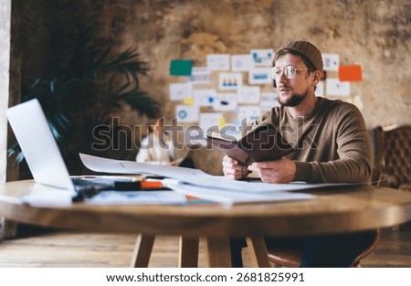 Focused businessman explains startup notes while holding notebook, developing digital concept in tech-oriented office filled with charts, diagrams and coworking energy.