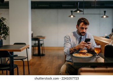 Focused businessman sitting alone in the local cafe, having a lunch break with a burger and beer and texting on his phone.
