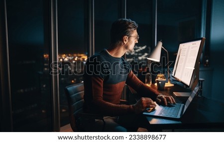 A focused businessman works late in a well-lit office, typing on his laptop. Surrounded by a busy workplace, he diligently focuses on a marketing plan, driven by ambition and dedication.