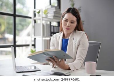 Focused businesswoman reviewing notes at bright office desk, surrounded by technology and paperwork, exuding confidence and determination - Powered by Shutterstock